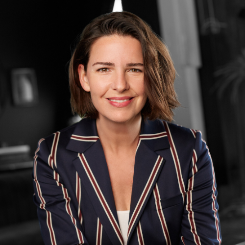 Melanie Love wearing a navy blazer with red and white stripes against a blurred indoor background
